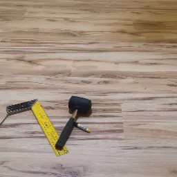 Fragment podłogi z paneli imitujących drewno w trakcie układania, widoczne narzędzia: gumowy młotek, kątownik, miarka i ołówek.