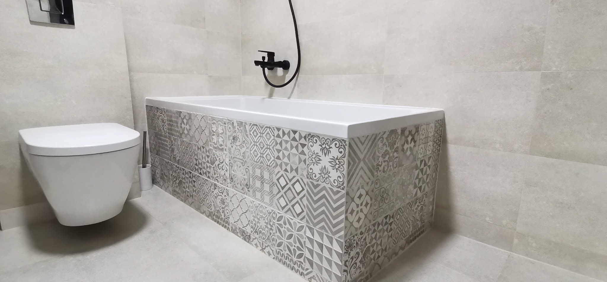 Nowoczesna łazienka w odcieniach szarości i bieli, z wiszącą toaletą i obudowaną wanną z dekoracyjnymi płytkami w geometryczne i kwiatowe wzory, czarna armatura.