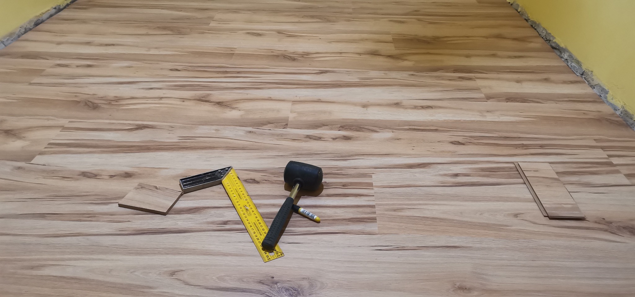 Fragment podłogi z paneli imitujących drewno w trakcie układania, widoczne narzędzia: gumowy młotek, kątownik, miarka i ołówek.