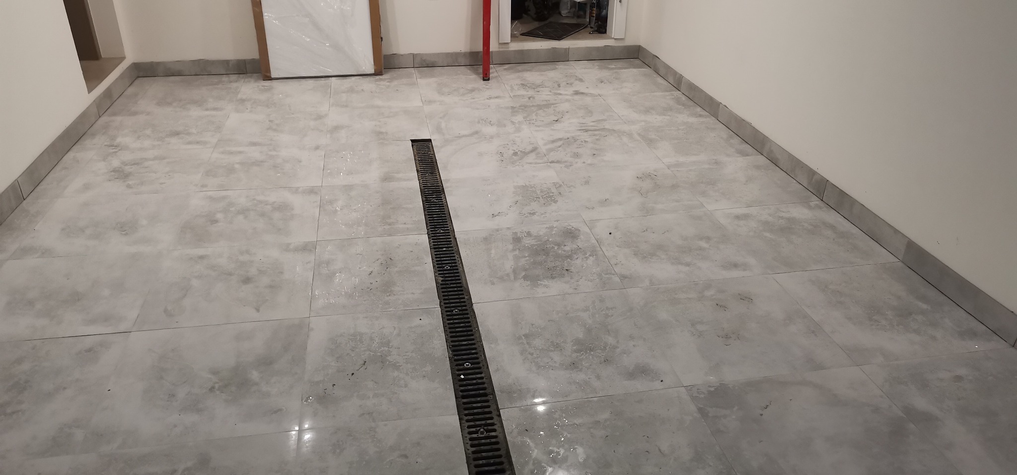 Wykończona podłoga w łazience z szarych płytek ceramicznych z odpływem liniowym, widoczna ściana z przypodłogowymi listwami