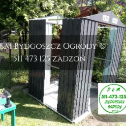 Montaż szarej, metalowej konstrukcji szopy ogrodowej w trakcie realizacji, widoczne narzędzia na zielonym stoliku, logo firmy w prawym dolnym rogu zdjęcia.