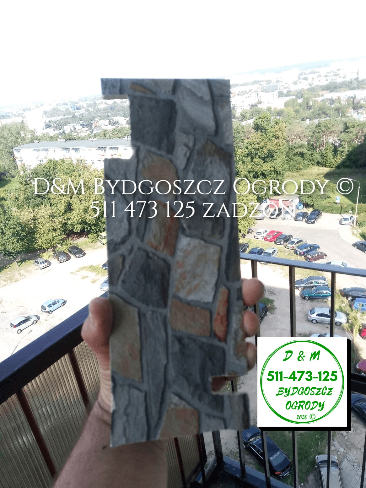 Próbka szarej okładziny kamiennej z elementami brązowymi, trzymana na balkonie z widokiem na miasto i parking.