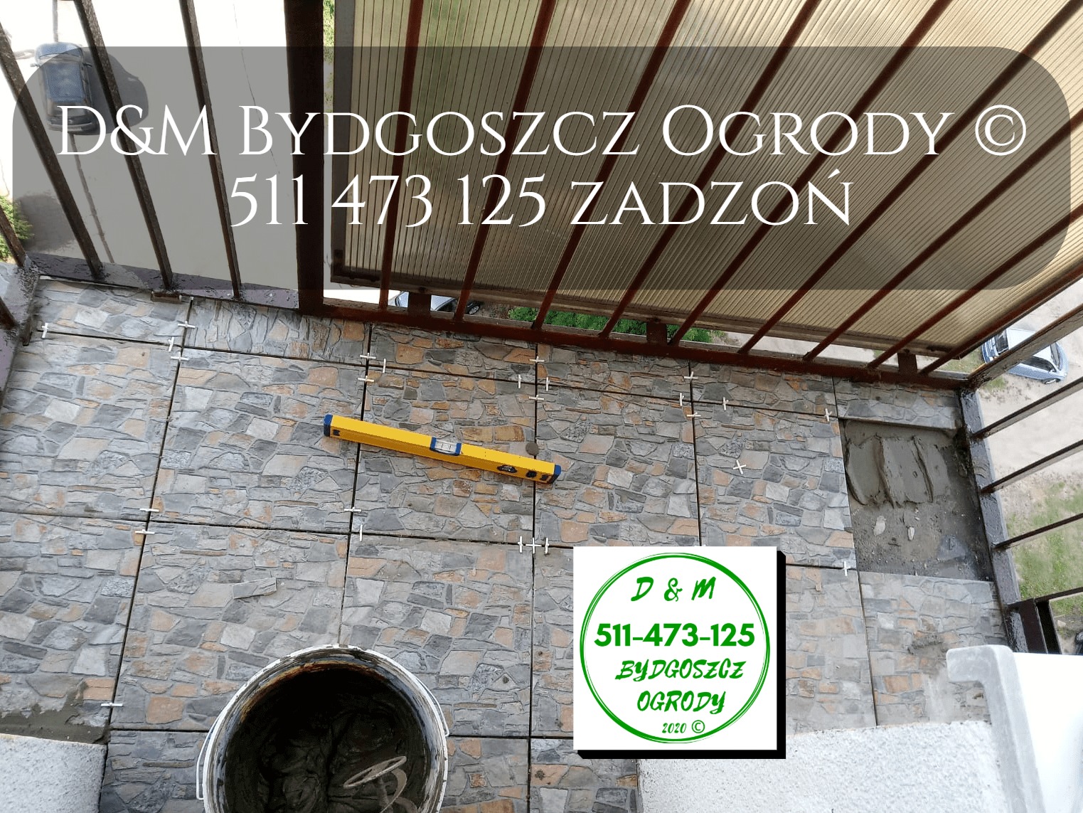 Układanie płytek imitujących kamień na zadaszonym tarasie, widoczny poziomica, wiadro z zaprawą i logo firmy D&M Bydgoszcz Ogrody