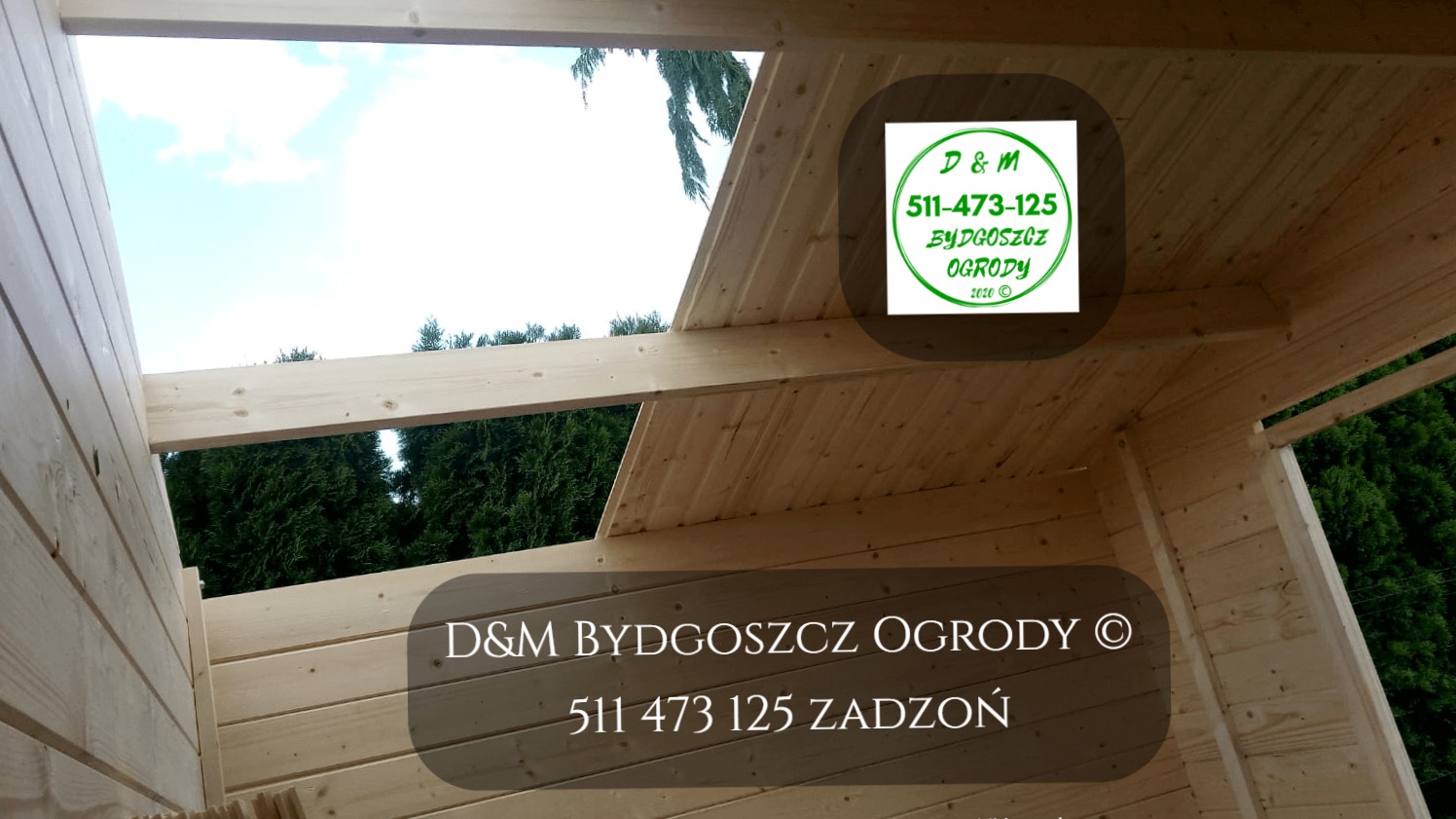 Wnętrze konstrukcji drewnianej w trakcie budowy, widoczne elementy dachu i ścian, z zewnątrz zieleń drzew, logo firmy D&M Bydgoszcz Ogrody naklejone na elementach konstrukcyjnych.