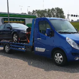 Niebieska laweta Renault Master z załadowanym ciemnym kombi Audi A3, zabezpieczonym pomarańczowymi pasami transportowymi, na parkingu przed myjnią samochodową.