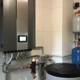 Nowoczesny srebrny moduł grzewczy z wyświetlaczem, podłączony do instalacji z dwoma zbiornikami wyrównawczymi (szarym i niebieskim) oraz zasobnikiem ciepłej wody w pomieszczeniu wyłożonym białymi...