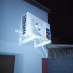 Biała jednostka zewnętrzna klimatyzatora zamontowana na ścianie budynku w słabym oświetleniu nocnym. Widoczne białe rurki prowadzące do wnętrza. Na boku urządzenia naklejka producenta.