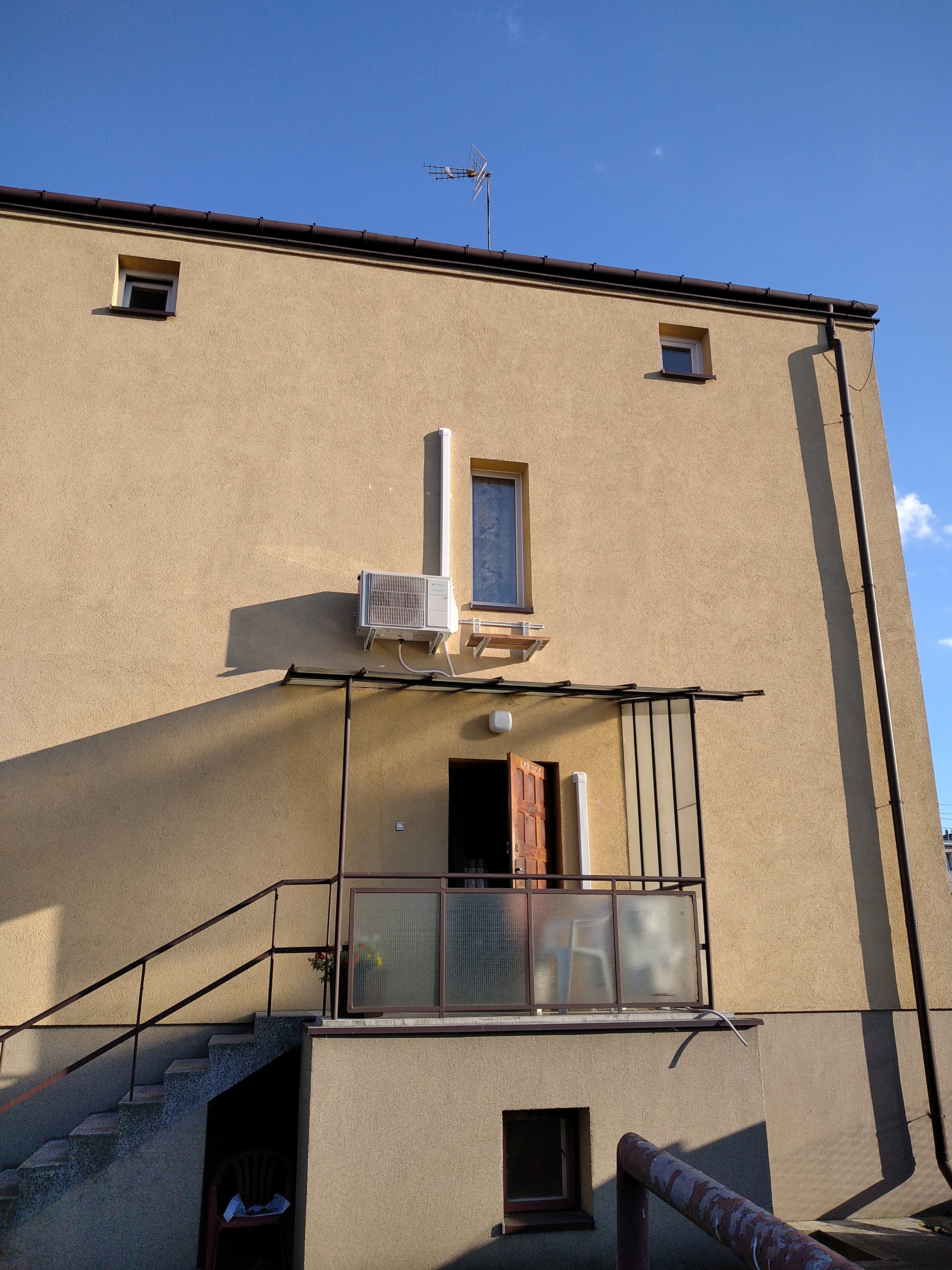 Zewnętrzna jednostka klimatyzacji zamontowana na elewacji budynku z otwartymi drzwiami balkonowymi i schodami wejściowymi.