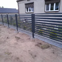 Nowoczesne, poziome ogrodzenie metalowe w kolorze grafitowym, osadzone na betonowej podmurówce, widoczne na tle domu z szarymi roletami zewnętrznymi, teren wokół ogrodzenia jeszcze niezagospodarowany.