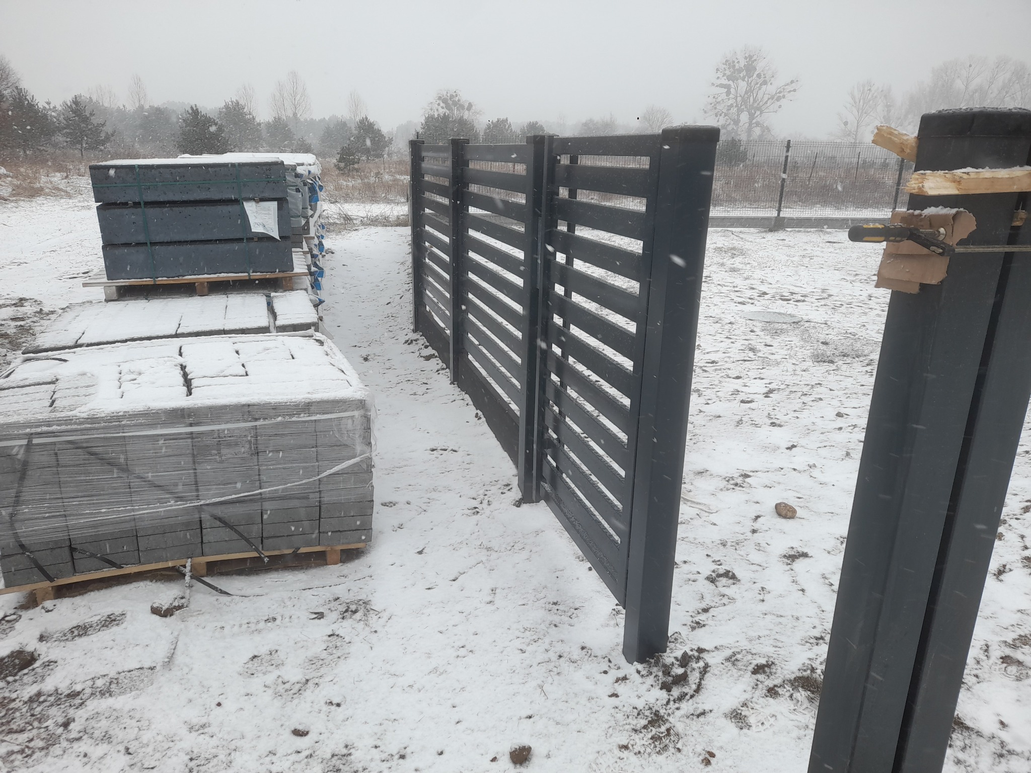 Częściowo zamontowane, poziome, szare ogrodzenie w zimowej scenerii. W tle stosy materiałów budowlanych i drzewa pod śniegiem. Widoczne narzędzia mocujące słupki.