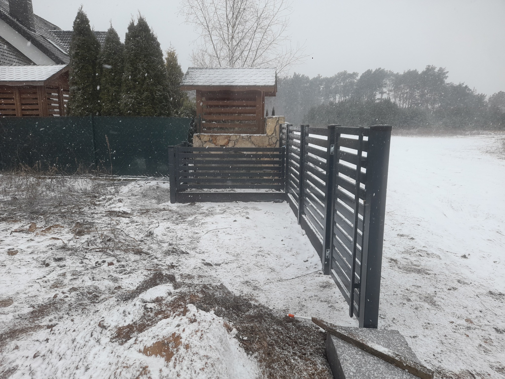 Montaż nowoczesnego, grafitowego ogrodzenia panelowego z poziomymi szczeblami w zimowej scenerii; widoczne narzędzia i elementy montażowe sugerują trwający proces instalacji.