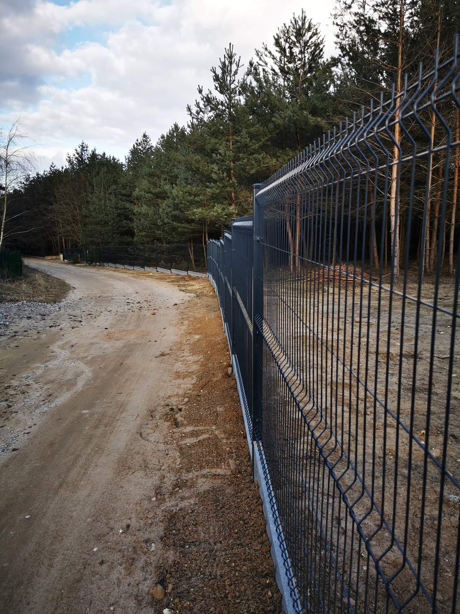 Ciemnoszare ogrodzenie z paneli drucianych na betonowej podmurówce, biegnące wzdłuż piaszczystej drogi w lesie, widok z boku.