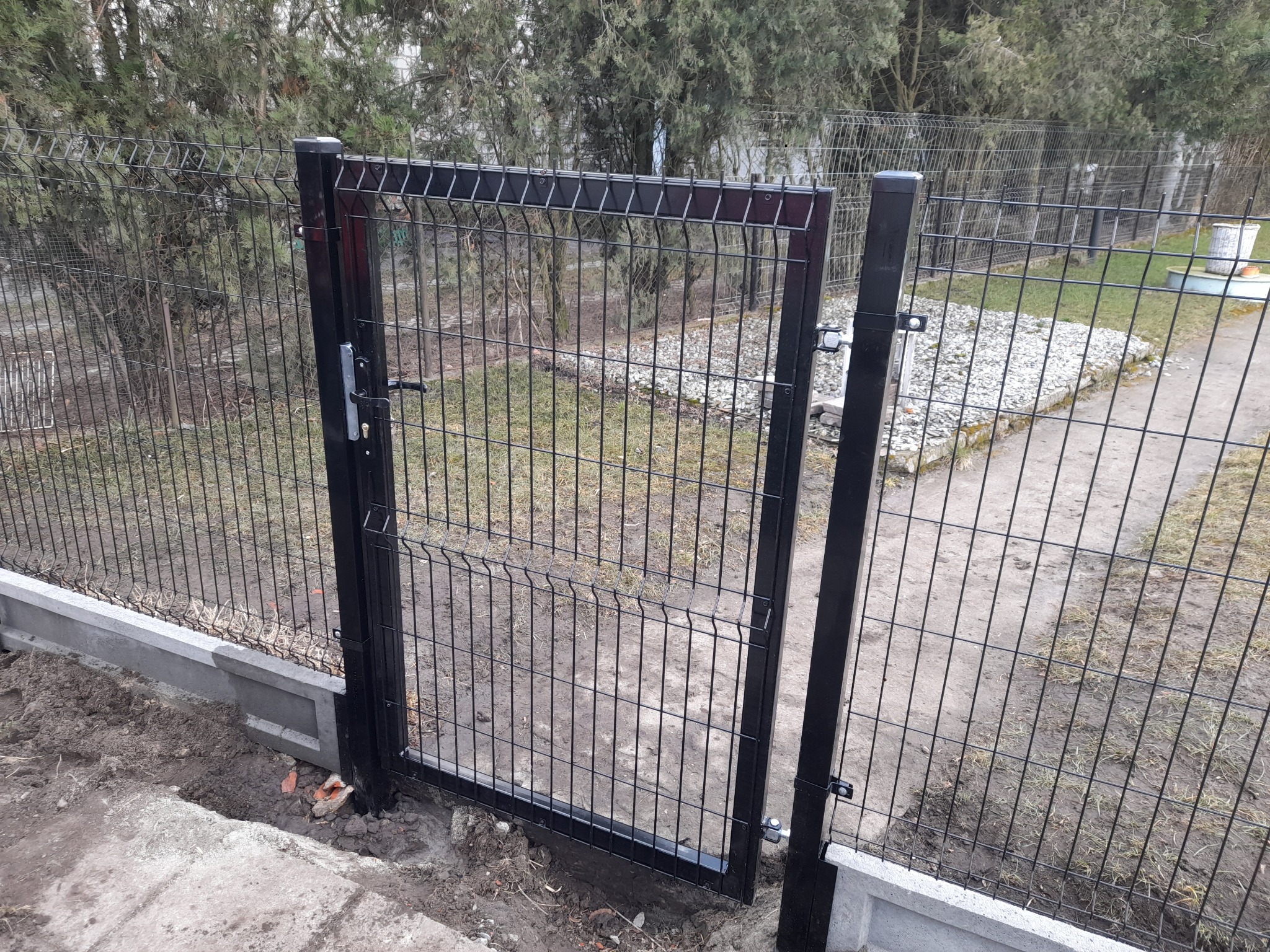 Otwarta furtka z czarnego, metalowego panelu ogrodzeniowego, zamontowana na betonowej podmurówce, z widocznym zamkiem i zawiasami, na tle zieleni.