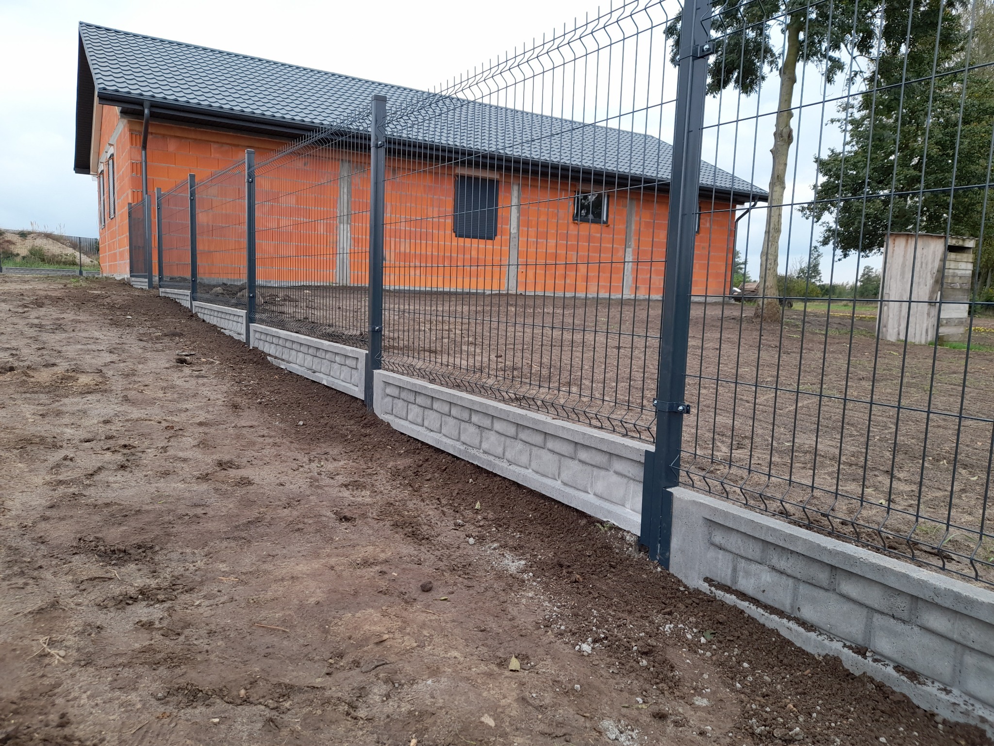 Szary panelowy system ogrodzeniowy z betonową podmurówką na nierównym terenie, widoczny budynek w trakcie budowy z cegły.
