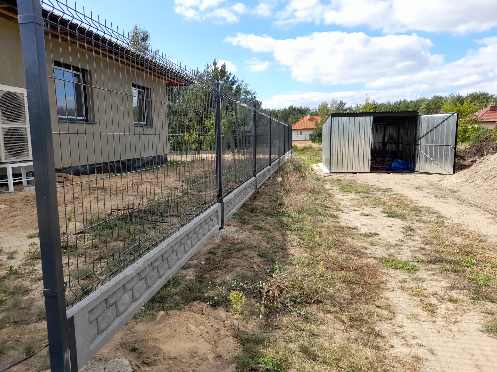 Nowoczesne, grafitowe ogrodzenie z siatki z podmurówką z betonowych bloczków, widok z boku na tle domu i metalowego garażu w słoneczny dzień.