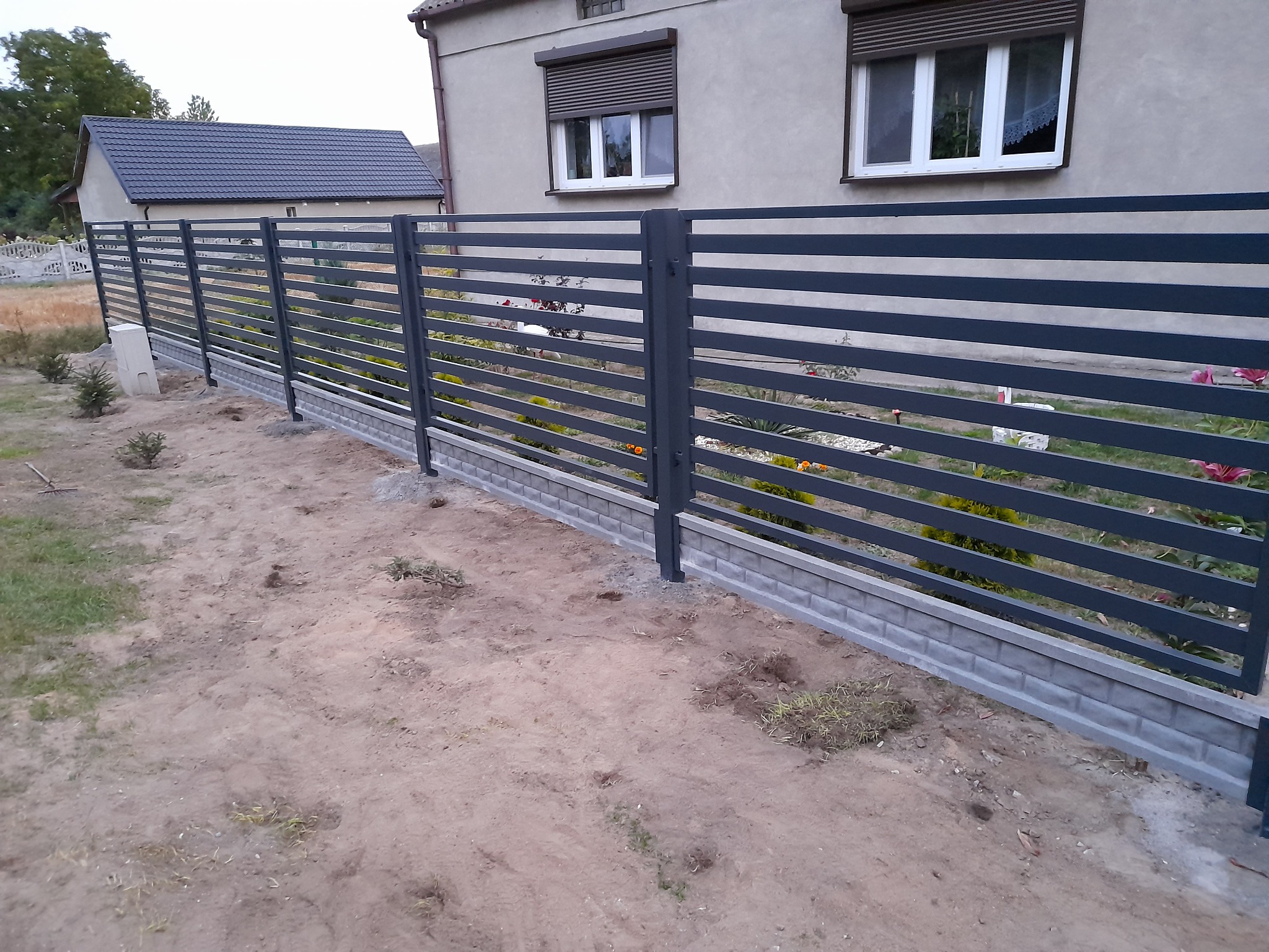 Nowoczesne, poziome ogrodzenie metalowe w kolorze grafitowym, osadzone na betonowej podmurówce, widoczne na tle domu z szarymi roletami zewnętrznymi, teren wokół ogrodzenia jeszcze niezagospodarowany.
