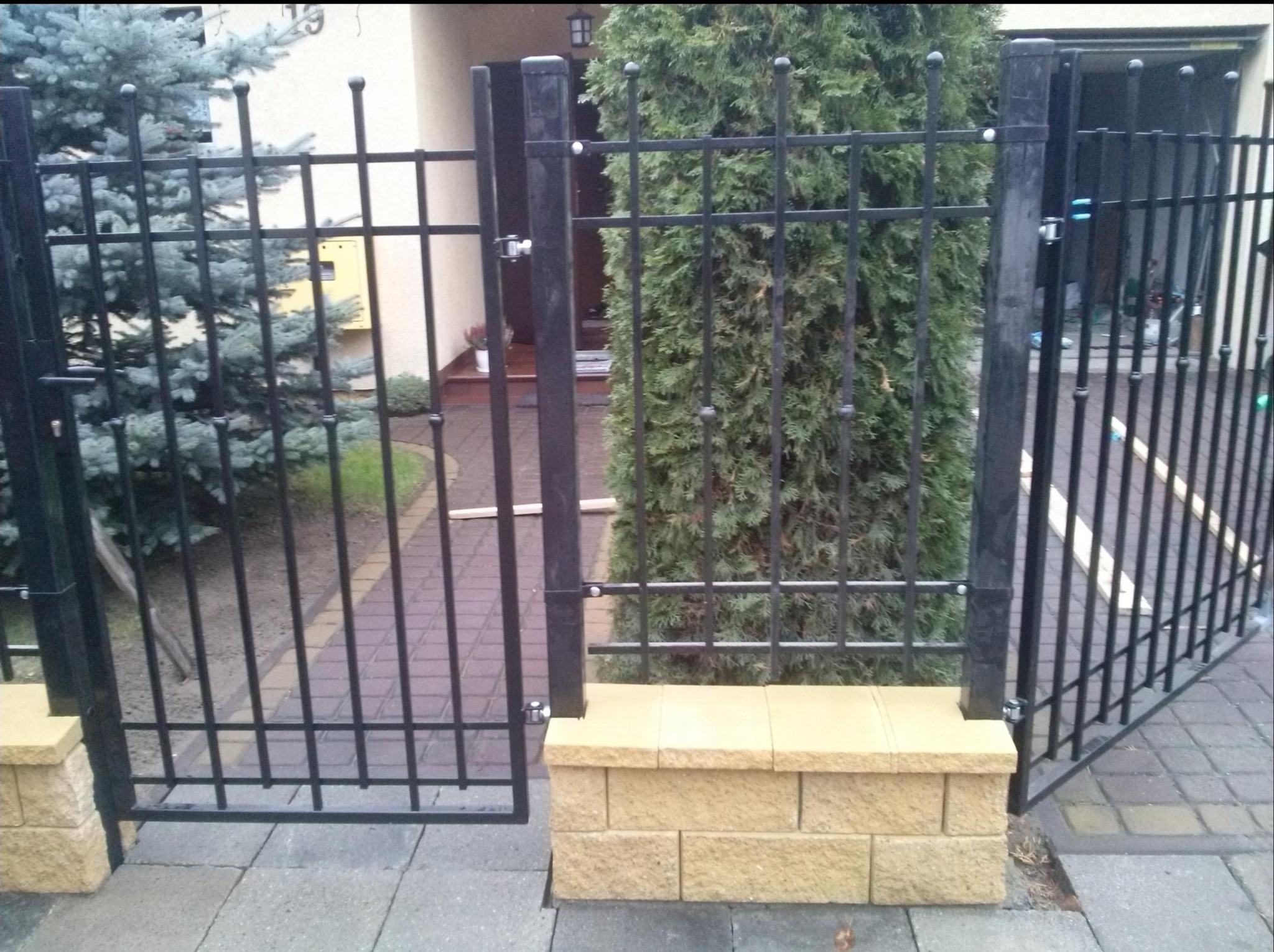 Czarna, metalowa brama dwuskrzydłowa z kulkami na szczycie, zamontowana na słupkach z żółtej kostki, widoczna z perspektywy chodnika z kostki brukowej.