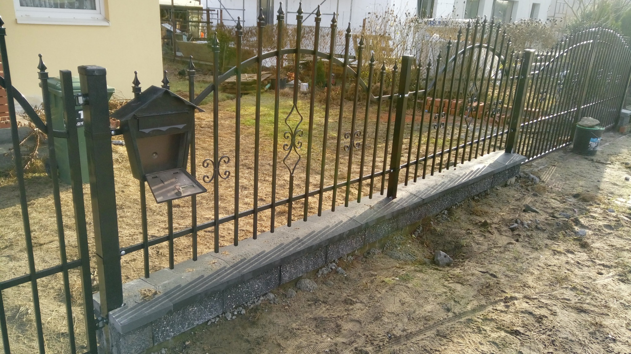 Kute, metalowe ogrodzenie w kolorze grafitowym z dekoracyjnymi elementami, osadzone na podmurówce z szarych bloczków betonowych; przy ogrodzeniu skrzynka pocztowa z otwartymi drzwiczkami.
