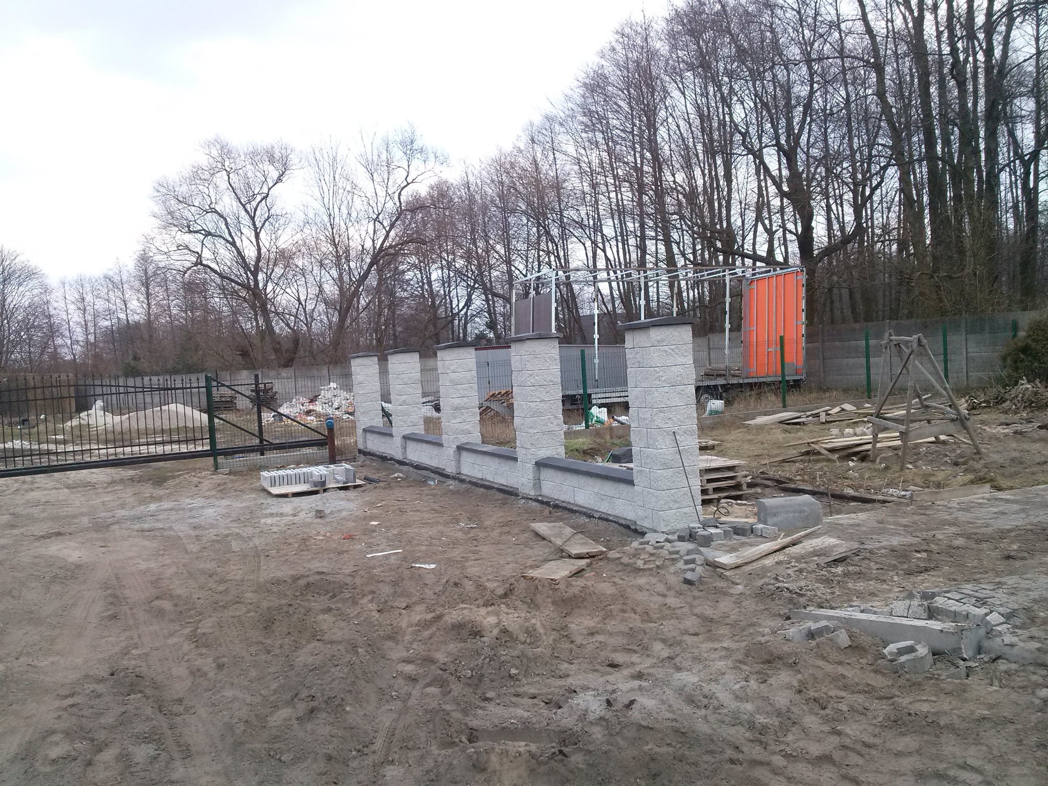 W trakcie budowy ogrodzenia z betonowych bloczków, widoczne słupki i podmurówka, teren budowy w trakcie prac ziemnych, w tle brama wjazdowa i drzewa bez liści.