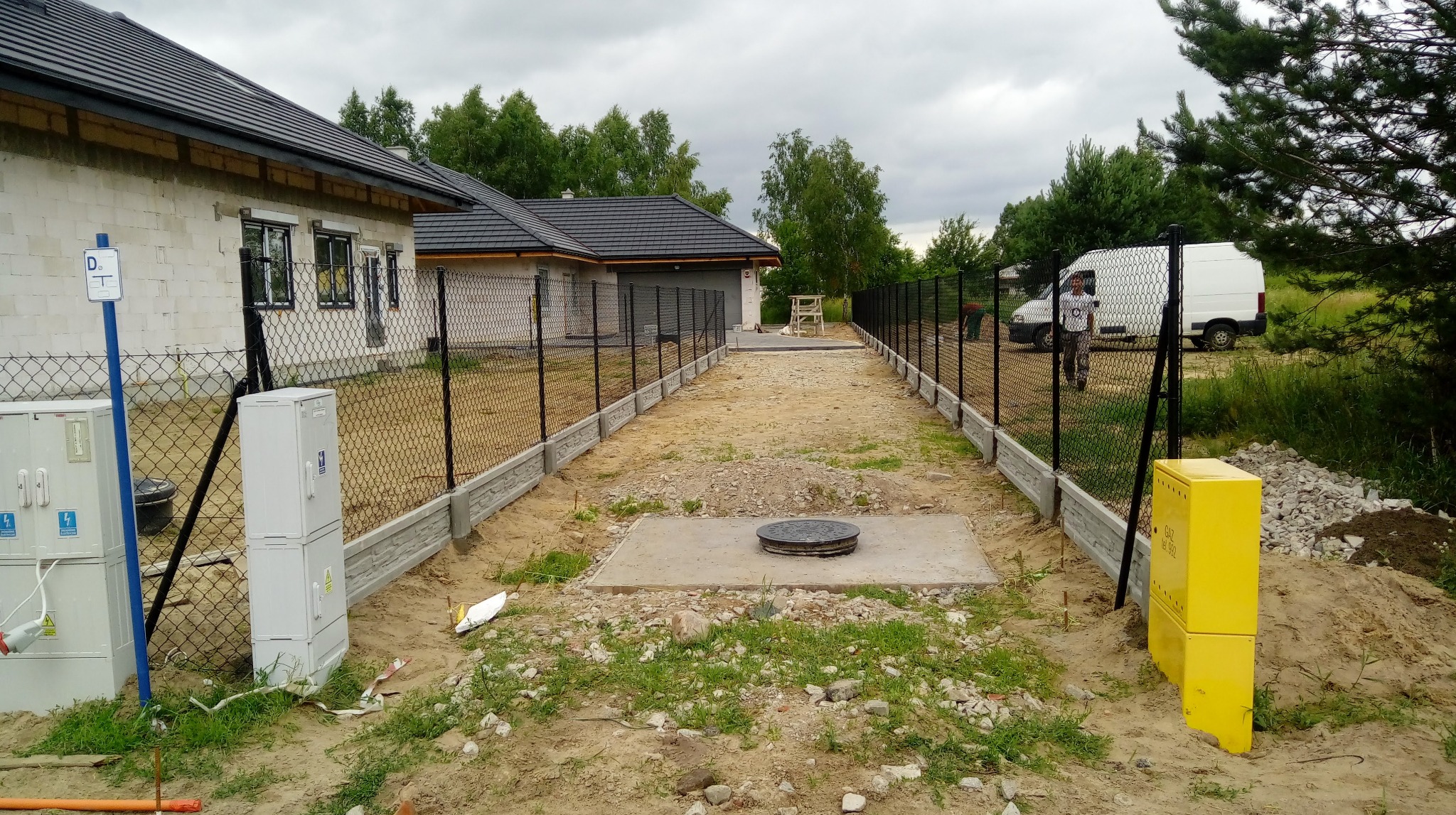 Nowe ogrodzenie z siatki z podmurówką betonową oddzielające posesje, widoczna szafka gazowa i skrzynki elektryczne, w tle dom w trakcie budowy i biały samochód dostawczy.