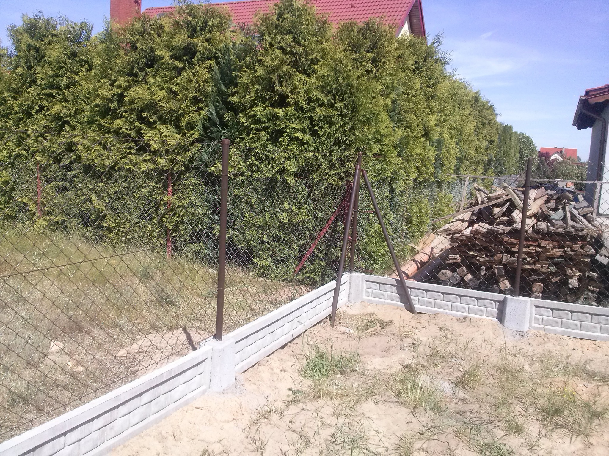 Wykop pod fundament ogrodzenia z siatki, widoczne betonowe prefabrykaty imitujące cegłę, stalowe słupki oraz pryzma pociętego drewna w tle.