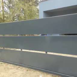 Nowoczesna przesuwna brama wjazdowa z poziomych paneli w kolorze grafitowym, częściowo owinięta folią ochronną, na tle domu i drzew.