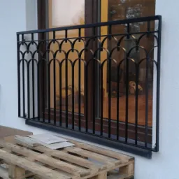 Czarna, metalowa balustrada okienna z elementami dekoracyjnymi w kształcie łuków, zamontowana na białej ścianie budynku w Brwinowie. W tle widoczne okno z refleksami i częściowo zasłonięta...