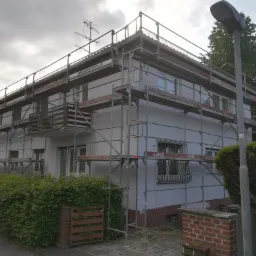 Dwupiętrowy dom jednorodzinny otoczony rusztowaniem, przygotowywany do prac elewacyjnych, widoczna antena na dachu i uliczna latarnia przed budynkiem.