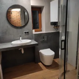 Nowoczesna łazienka z szarą płytką imitującą beton, drewnopodobną podłogą, okrągłym lustrem odbijającym ceglaną ścianę, wiszącą toaletą i kabiną prysznicową z czarnymi profilami.