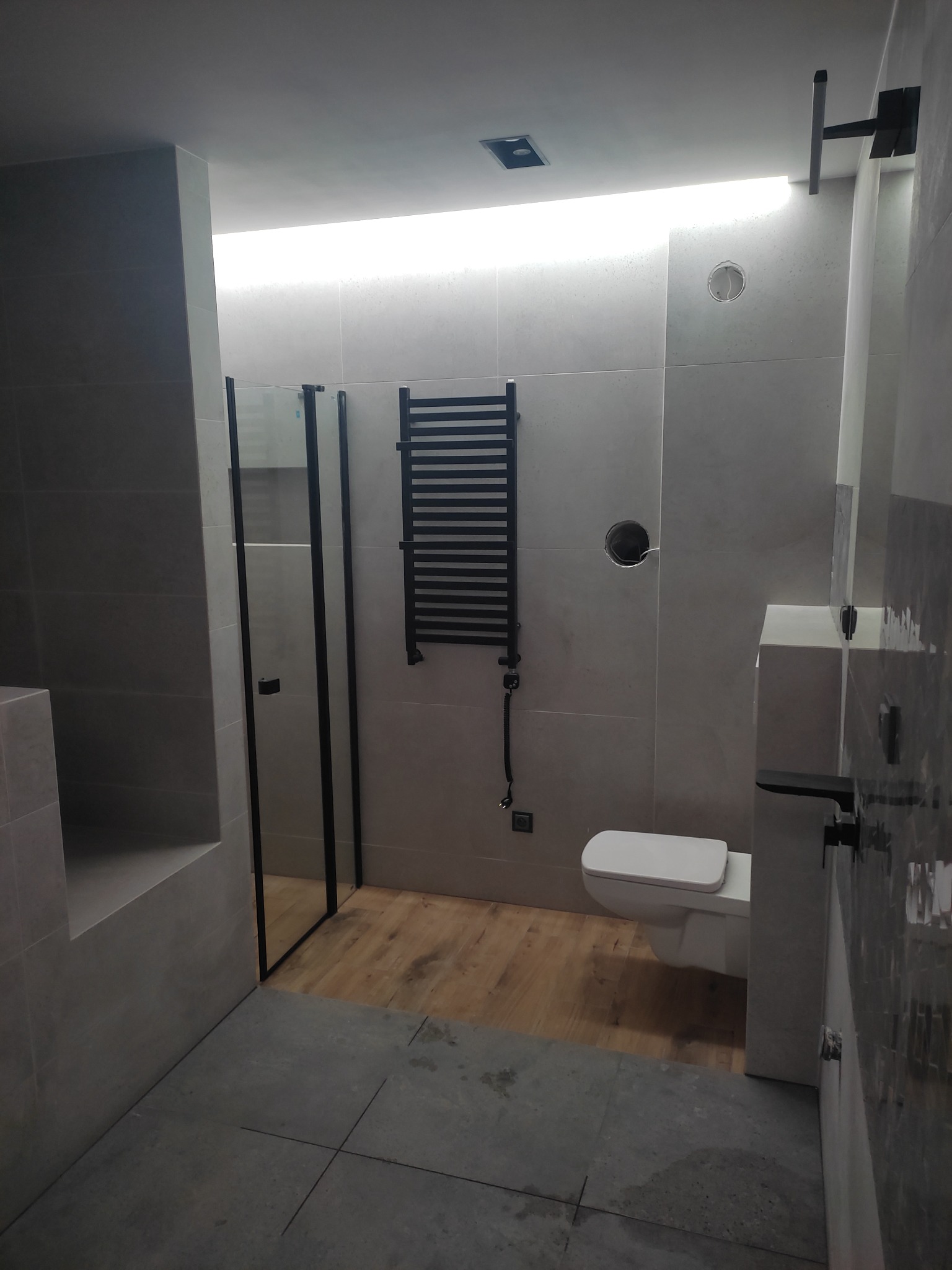 Nowoczesna łazienka w szarościach z drewnianą podłogą, kabina prysznicowa z czarnymi ramami, grzejnik drabinkowy i podświetlenie LED.