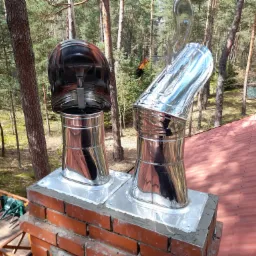 Dwa kominy ze stali nierdzewnej, jeden z czarną nasadą, zamontowane na ceglanym kominie na tle lasu i dachu z czerwonej dachówki.