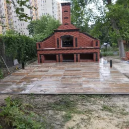 Wykończony taras z płytek imitujących drewno, z grillem murowanym z czerwonej cegły w tle, w trakcie deszczu, widoczne elementy montażowe ogrodzenia.