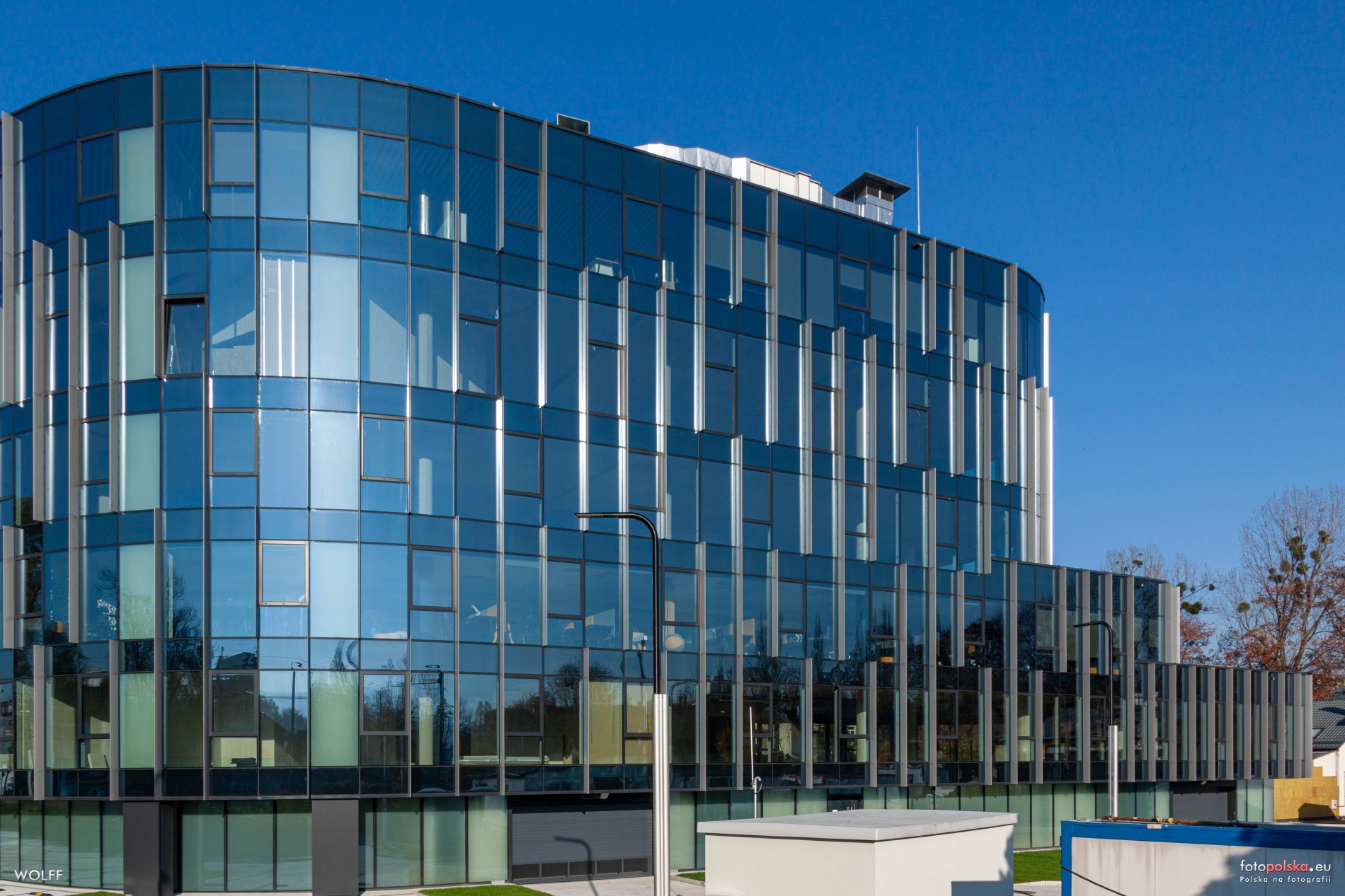 Nowoczesny biurowiec z fasadą ze szkła refleksyjnego, odbijającego błękit nieba, z widocznymi elementami konstrukcyjnymi i oświetleniem zewnętrznym.