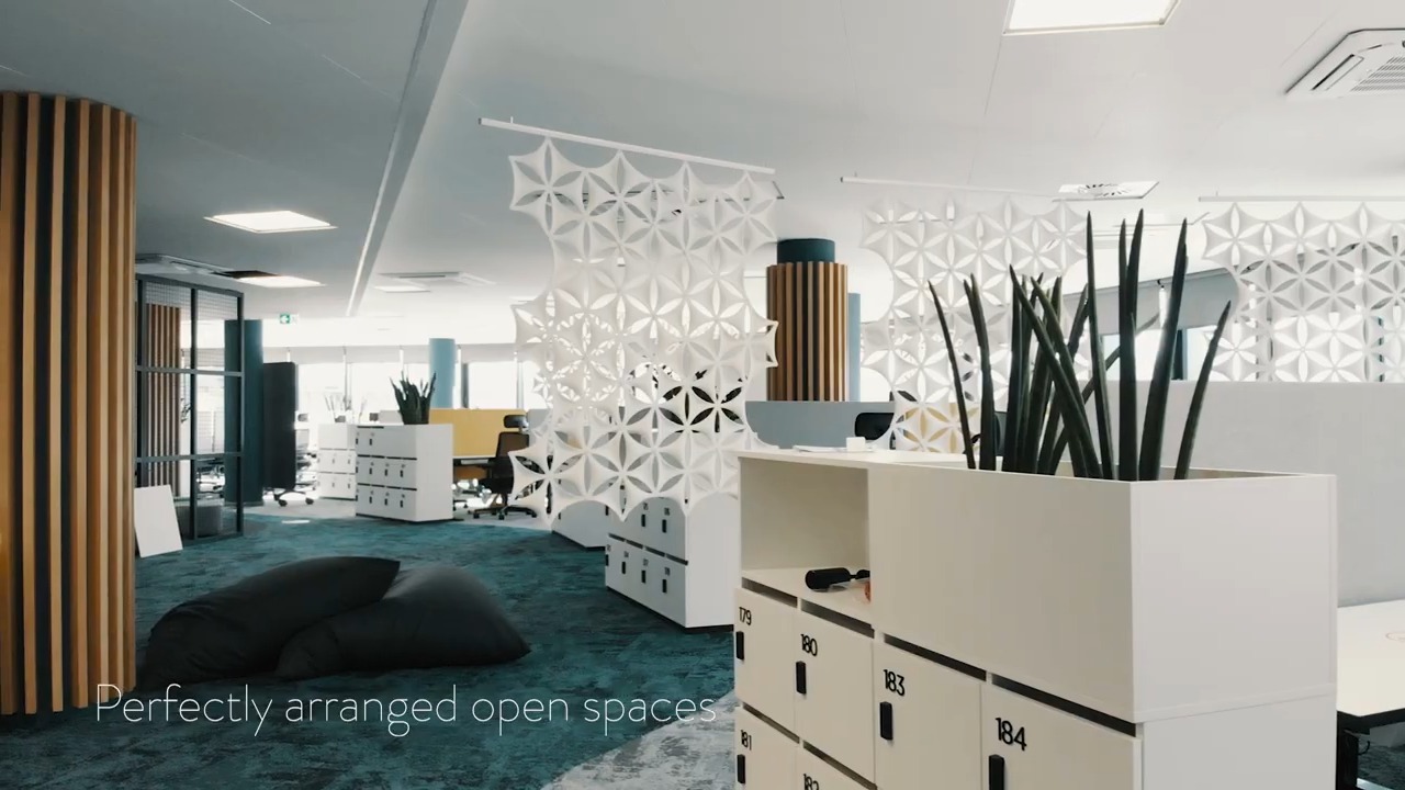 Nowoczesna aranżacja biura z białymi ażurowymi ściankami działowymi, roślinami w donicach na szafkach pracowniczych, drewnianymi elementami dekoracyjnymi i workami do siedzenia na turkusowej...