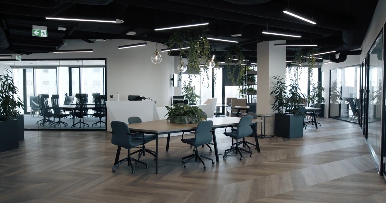 Jasne, przestronne biuro z podłogą w jodełkę, stołem konferencyjnym z szarymi krzesłami, szklanymi ścianami działowymi i roślinami doniczkowymi, oświetlone nowoczesnymi lampami sufitowymi...