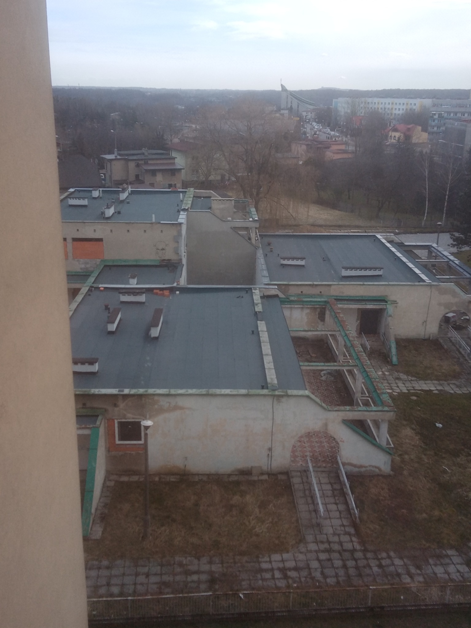 Widok z góry na płaskie dachy budynku w trakcie remontu, widoczne kominy wentylacyjne, częściowo zdemontowane elementy konstrukcyjne i otoczenie miejskie w tle.