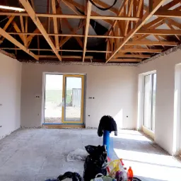 Wnętrze domu w trakcie prac wykończeniowych, ściany po nałożeniu gładzi, widoczna drewniana konstrukcja dachu, okna z żółtymi ramami, na podłodze resztki materiałów budowlanych i śmieci.