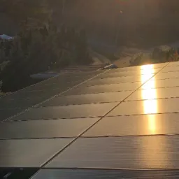 Panel słoneczny na dachu w promieniach zachodzącego słońca, z widocznym otoczeniem drzew i drogi