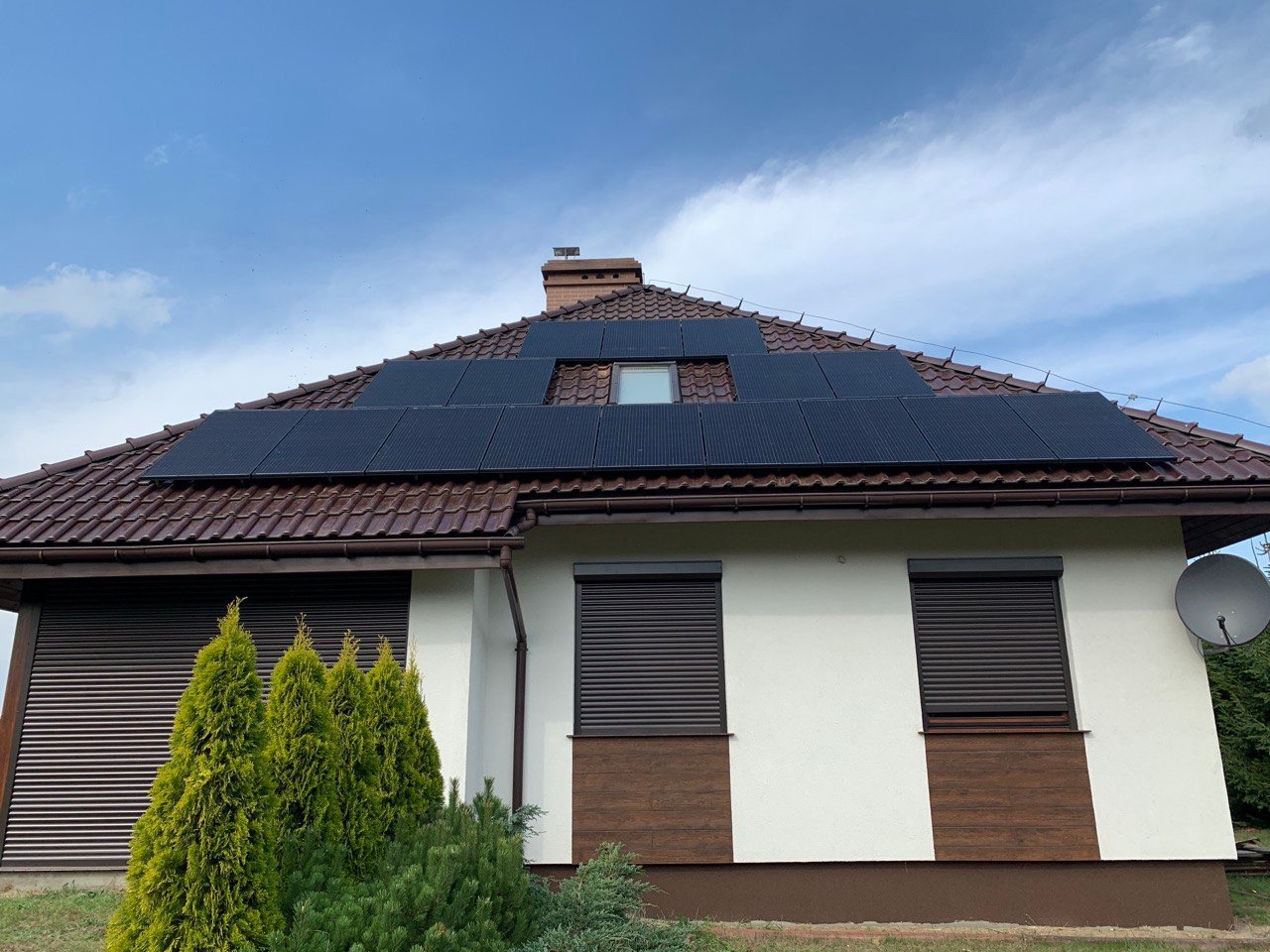 Dom jednorodzinny z brązowym dachem pokrytym panelami słonecznymi, widoczne rolety zewnętrzne i antena satelitarna.