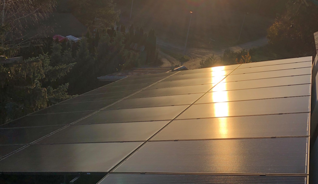 Panel słoneczny na dachu w promieniach zachodzącego słońca, z widocznym otoczeniem drzew i drogi