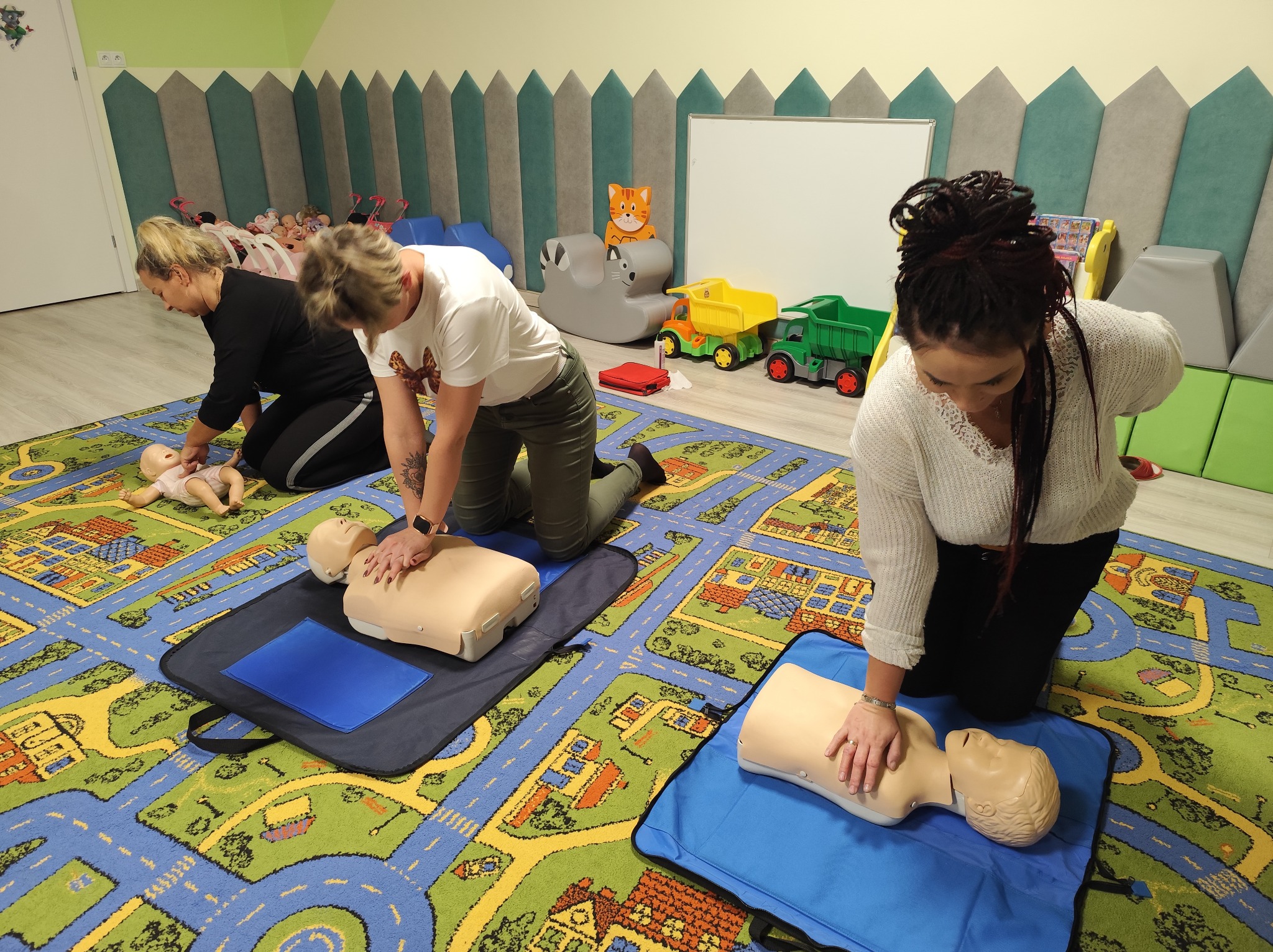 Szkolenie z resuscytacji krążeniowo-oddechowej na manekinach w sali z kolorowym dywanem i zabawkami w tle. Uczestniczki ćwiczą pod okiem instruktora.