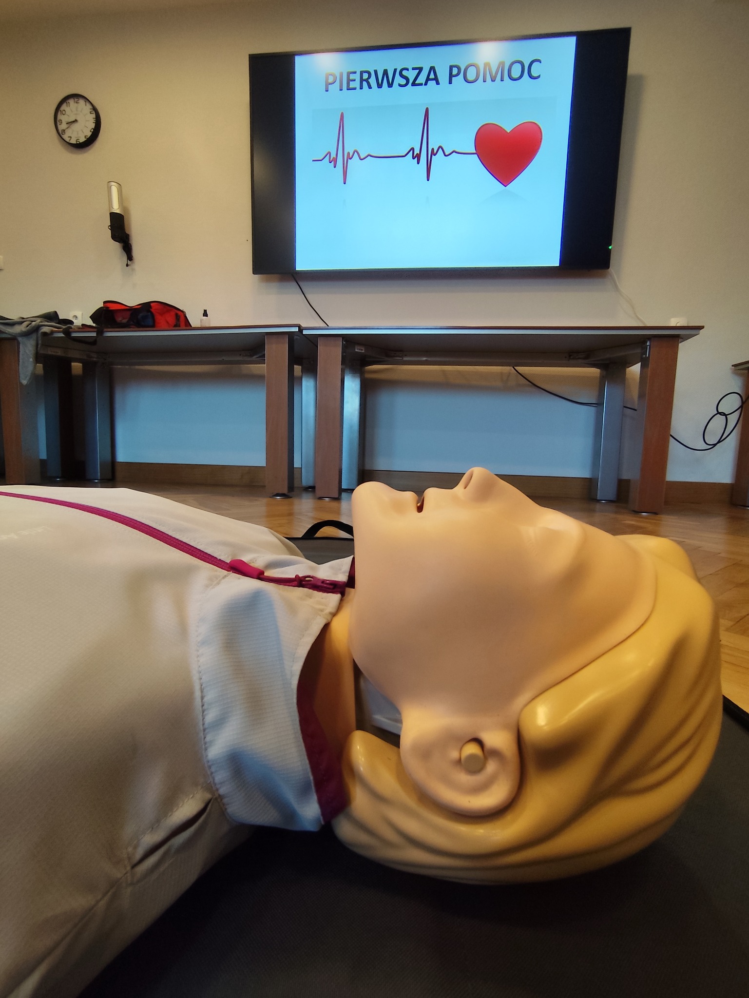 Manekin do ćwiczeń RKO leży na macie, w tle ekran z napisem 'Pierwsza Pomoc' i symbolem EKG. Sala szkoleniowa z drewnianą podłogą i stołami.