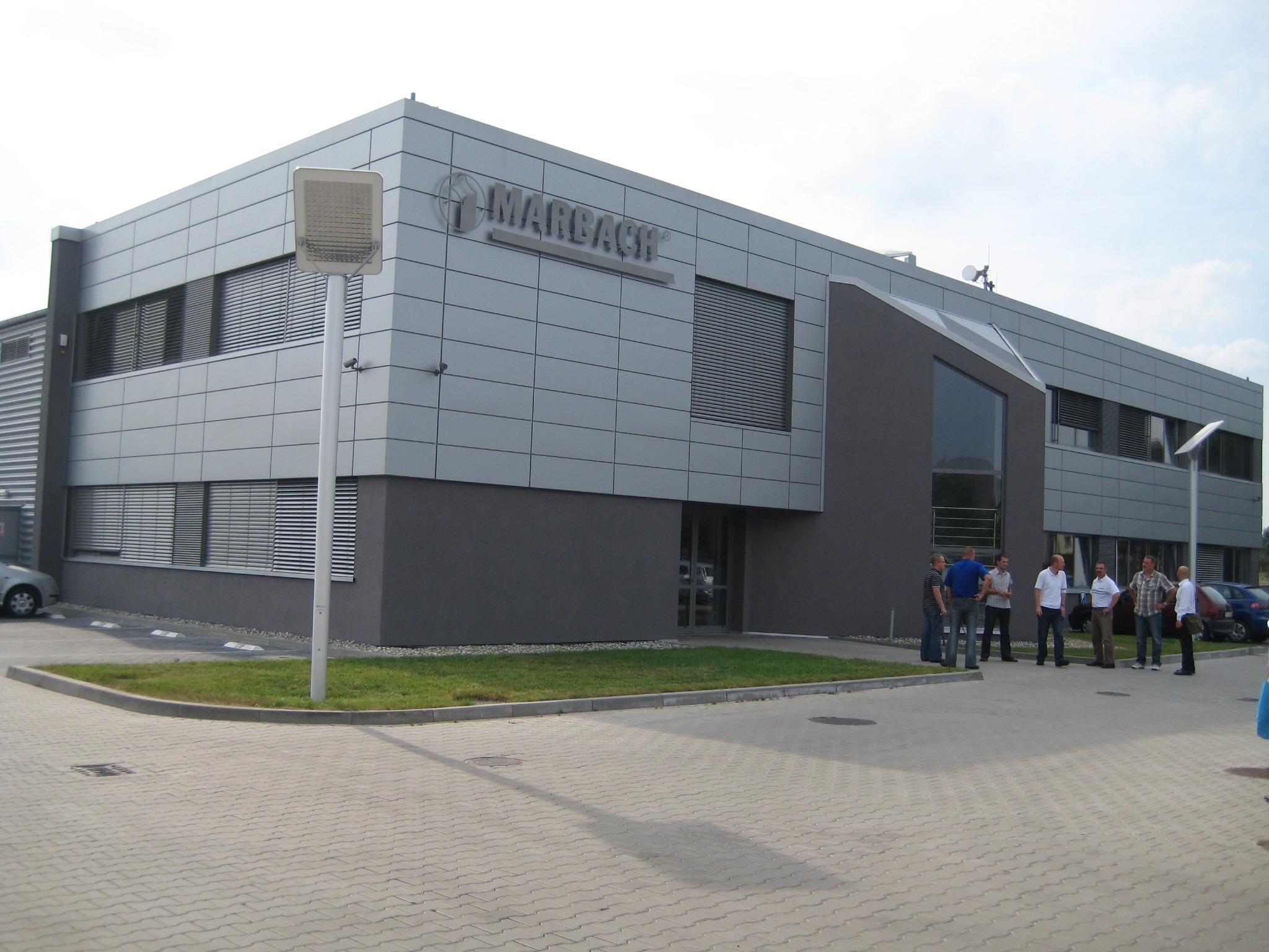 Nowoczesny budynek firmy Marbach z szarej blachy trapezowej i logo, grupa mężczyzn przed wejściem, oświetlenie zewnętrzne.