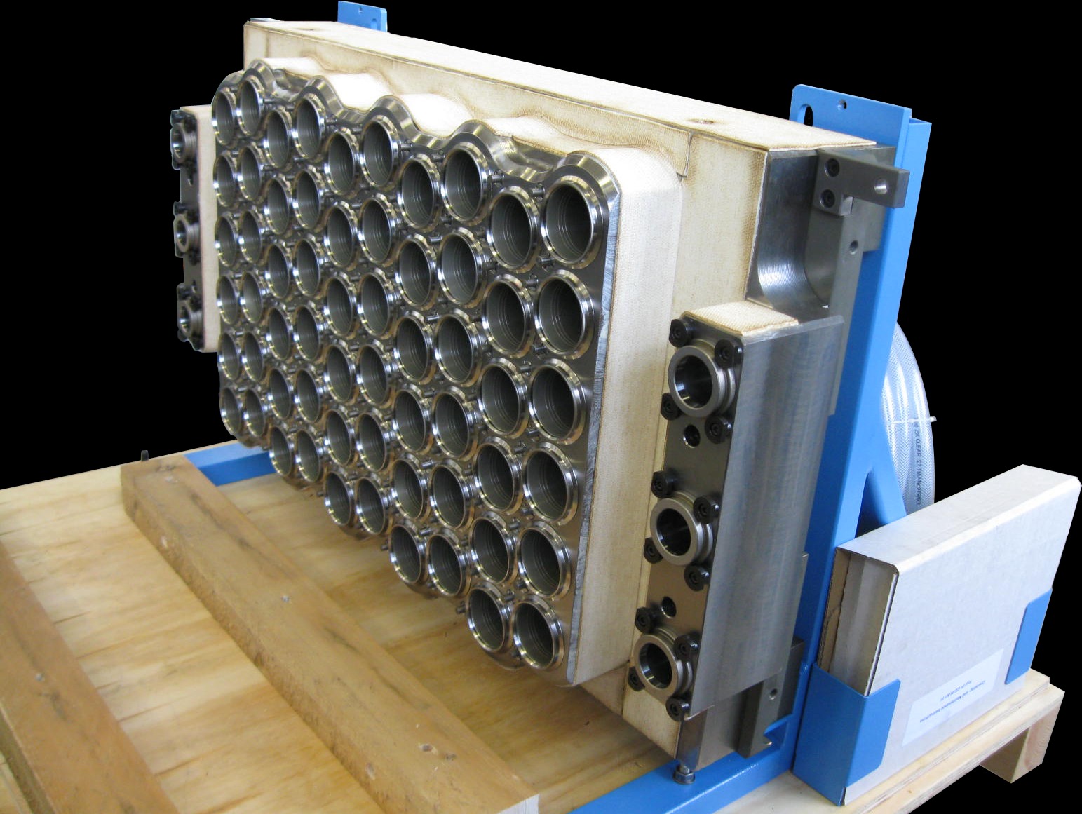 Metalowy blok z wieloma okrągłymi otworami, zamontowany na niebieskiej ramie, spoczywający na drewnianej palecie. Widoczne ślady obróbki i elementy mocujące.