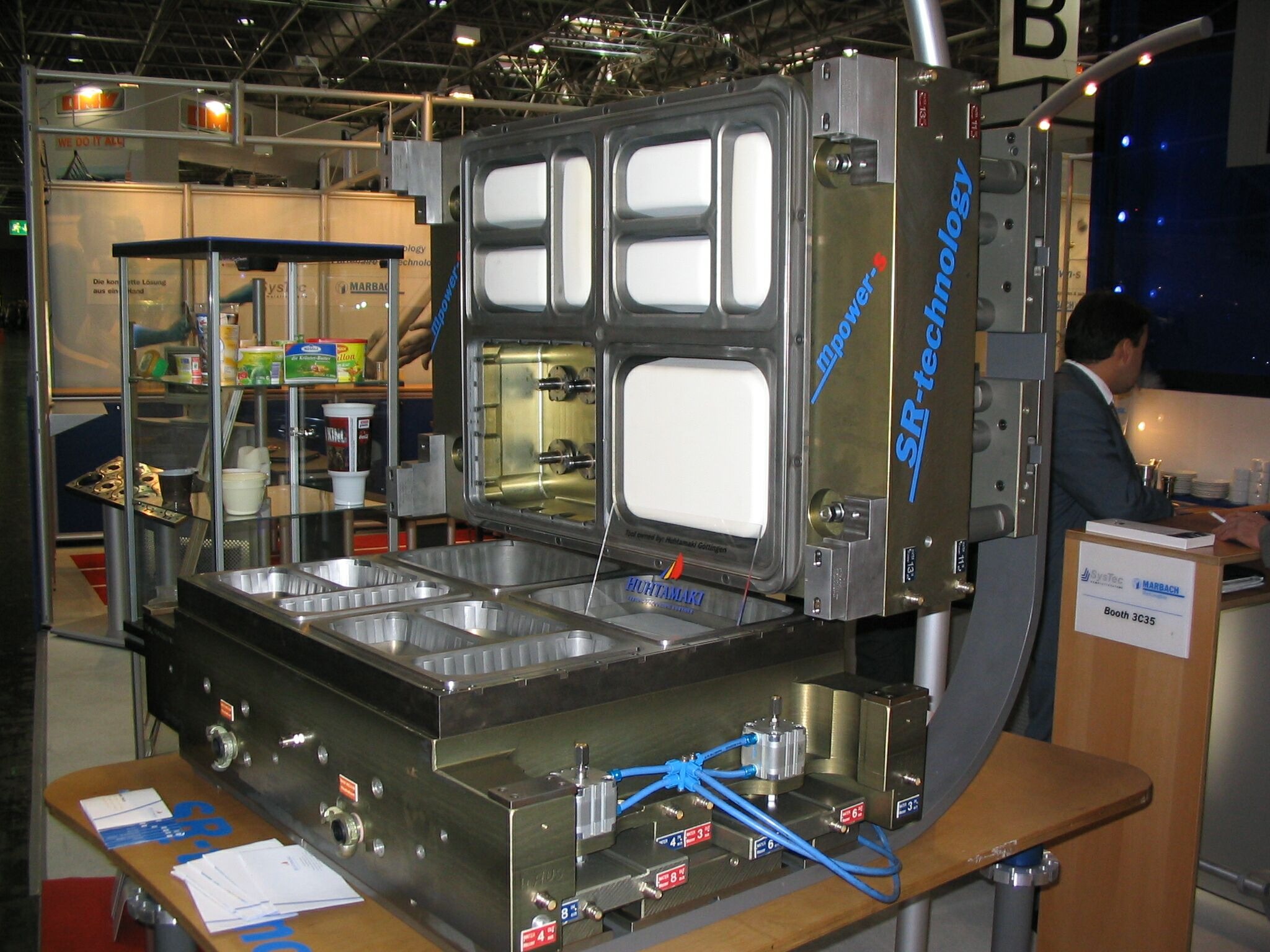 Precyzyjna maszyna do formowania tworzyw sztucznych z otwartą formą, widoczne elementy sterujące i niebieskie przewody pneumatyczne, prezentowana na stoisku targowym z logo Huhtamaki na wkładce formy.