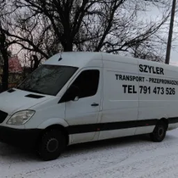 Biały samochód dostawczy Mercedes-Benz Sprinter z napisem 'SZYLER TRANSPORT - PRZEPROWADZKI TEL 791 473 526' zaparkowany na zaśnieżonym placu w pochmurny zimowy dzień.