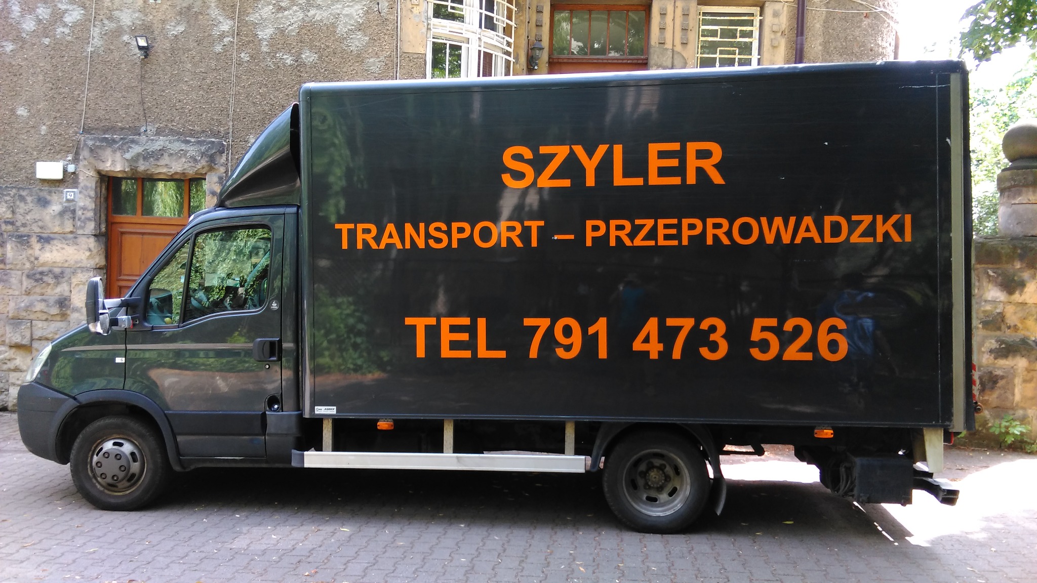 Czarny samochód dostawczy firmy transportowej z pomarańczowym napisem 'SZYLER TRANSPORT - PRZEPROWADZKI TEL 791 473 526' zaparkowany przed budynkiem z kamienia.
