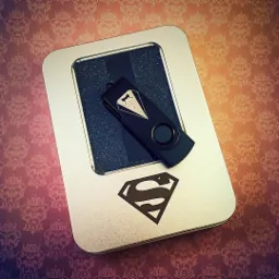 Czarny pendrive z nadrukiem smokingu leżący na srebrnym pudełku z logo Supermana, na tle wzorzystej tapety.