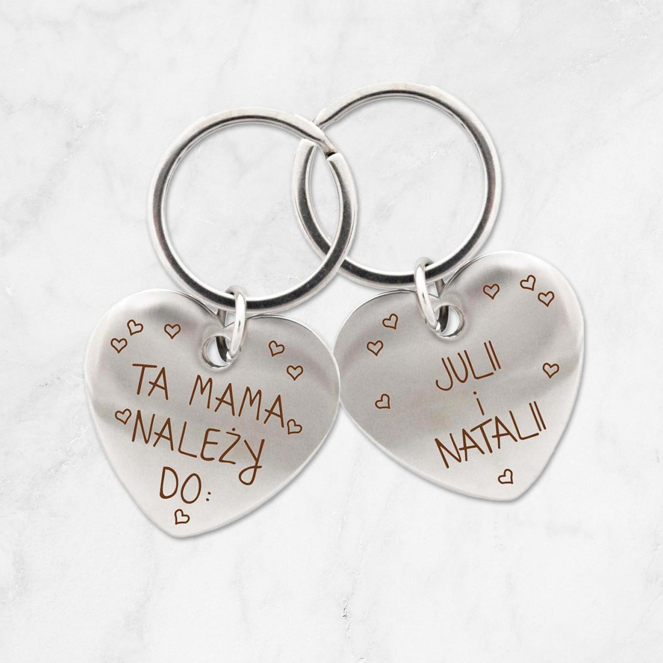Dwa srebrne breloczki w kształcie serc z grawerowanymi napisami: 'Ta Mama należy do:' i 'JULII i NATALII', otoczone małymi serduszkami, zawieszone na srebrnych kółkach.