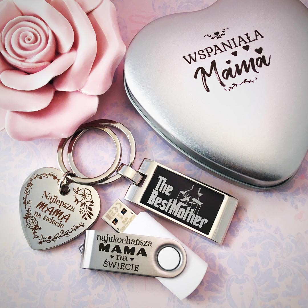 Srebrne pudełko w kształcie serca z napisem 'Wspaniała Mama', breloczek w kształcie serca z grawerem 'Najlepsza Mama na świecie' oraz dwa pendrive'y z napisami dedykowanymi dla mamy, leżące obok...